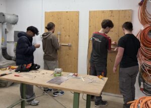Mehrere Jugendliche arbeiten in einem Werkraum an elektrischen Installationsübungen mit Werkzeug und Kabeln.